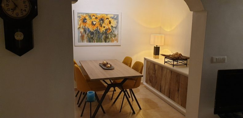 Eetkamer Huiselijke eetruimte met houten tafel, gele stoelen en een schilderij van zonnebloemen.